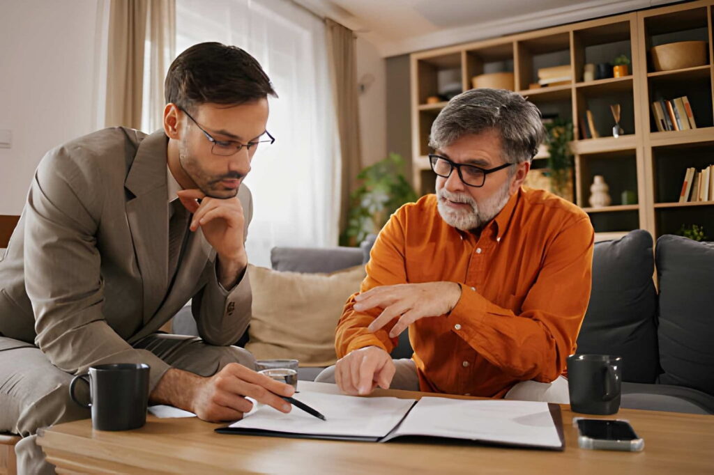 Two men sitting together at a table, using a laptop and a pen for their discussion or project.